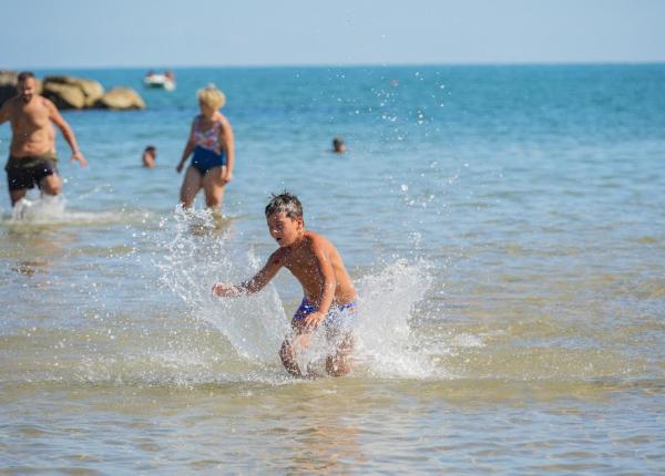 Kind spielt im Meer mit Wasserspritzern, Erwachsene im Hintergrund.